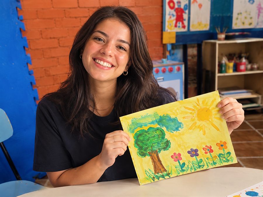 Jovem sorrindo mostrando um desenho colorido numa sala do Cantinho