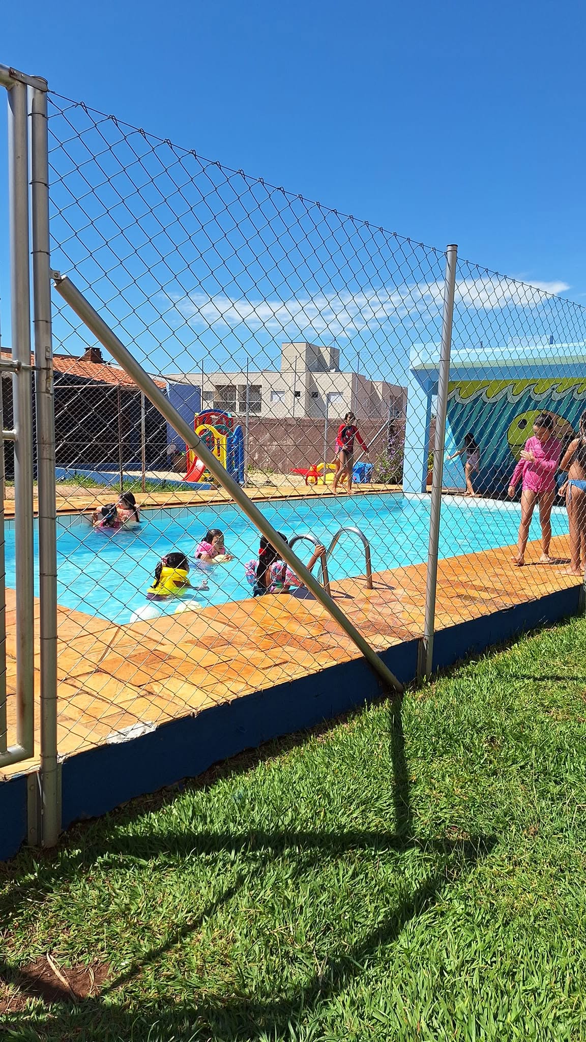 Piscina cercada por grade de proteção, com parquinho ao fundo